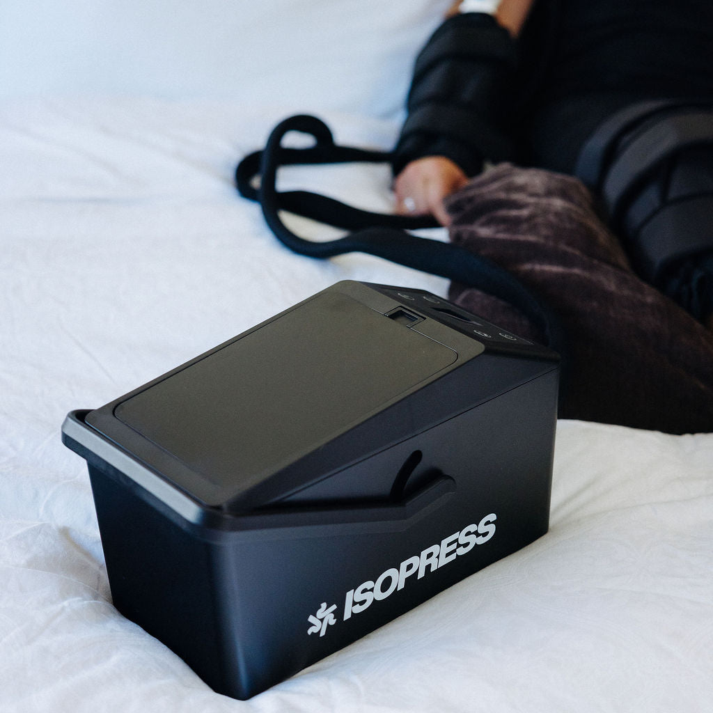 Black ISOPRESS device on a white surface with a person lying down in the background.