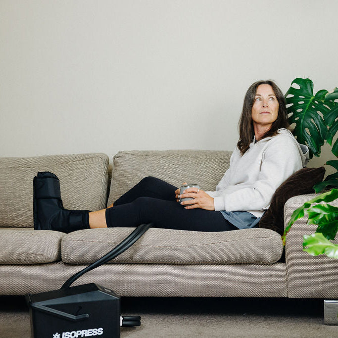 Woman sitting on a couch with a plant and a coffee cup, using a black compression device labeled 'Isopress'.