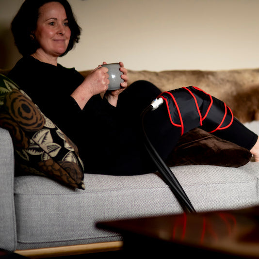 Woman sitting on a couch with a cold compression cuff on her knee, holding a cup, in a cozy living room setting.
